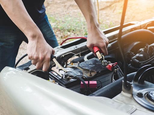 A man standing over the engine area of a vehicle with his hands placed on the ends of two jumper cables as he attaches it to the terminals on a car battery.