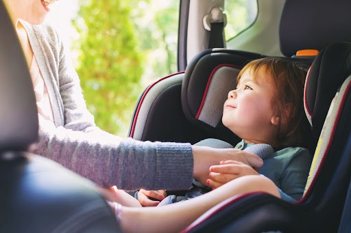 Child sitting in a car seat within a vehicle with its mother reaching into the car and buckling the child.