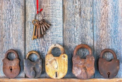 Five vintage padlocks in various colors leaning against a weathered wall. Above the locks, a ring of keys is hanging on a ribbon.