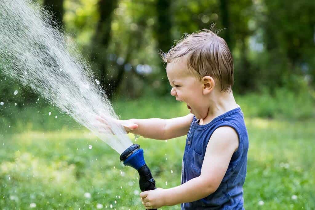 Young blonde child in a blue tank top holding a garden hose with water spraying out of it and putting their hand into the water stream.