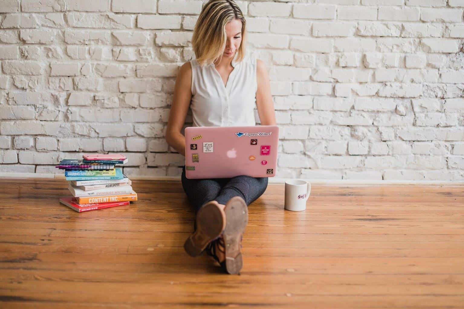 Blonde sitting on the floor with their back against a white brick wall typing on a laptop.