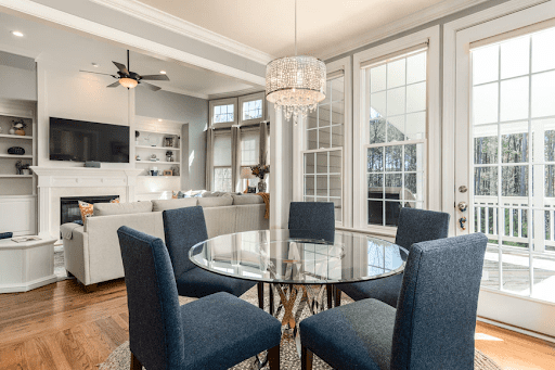 A photo of a bright, modern home. The shot is from the dining room into the kitchen and highlights beautiful, airy windows full of sun.