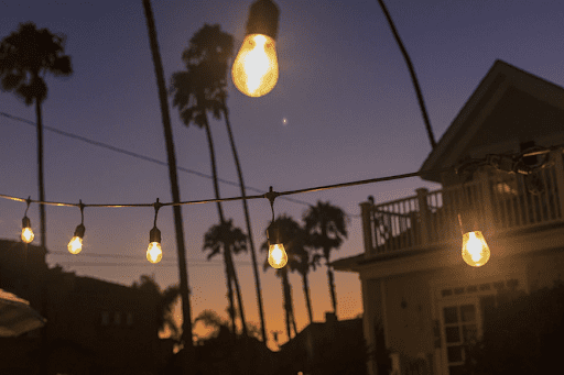 Photo of dusk where the sky is getting dark, but the horizon is filled with the sunset’s glow. In the image is a beach home with towering palm trees and outdoor hanging lights.