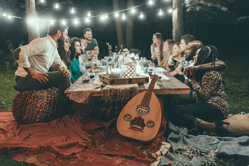 A photo of many friends sitting on the ground outside around a short table. There’s a guitar and the table is decorated with drinks, food, and other festive items.